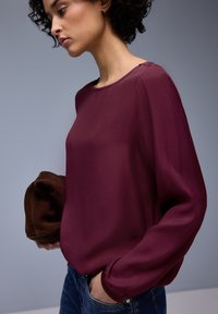 Blouse bordeaux en tissu léger, dotée d'un col rond et de manches longues légèrement bouffantes. Associée à un jean en denim.