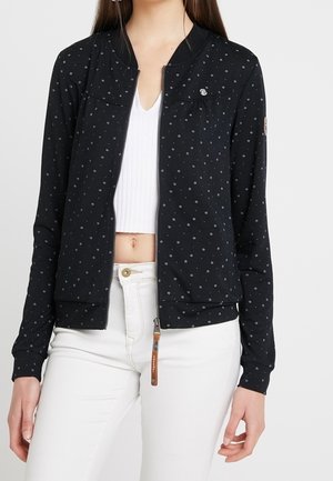 Kobieta ubrana w czarną kurtkę zapinaną na suwak z małymi białymi wzorami w kształcie gwiazdek, biały krótki top oraz białe spodnie z wysokim stanem.