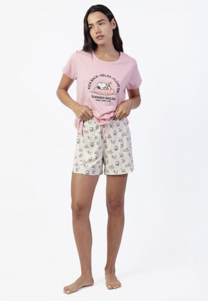 Mujer con camiseta rosa temática de Snoopy con texto "Kick Back, Relax, Float On" y pantalones cortos a juego con estampado del personaje, de pie descalza.