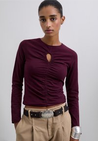 Burgundy long-sleeve top with a gathered center and keyhole detail, paired with tan trousers and a black belt featuring a silver buckle.