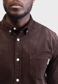 Camisa de pana marrón con cuello abotonado, botones blancos y un bolsillo en el pecho izquierdo. Textura suave con sutiles estrías.