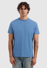 Blauwe katoenen t-shirt met een ronde hals en korte mouwen. Heeft een kleine bedrukte tekst "PURE PATH" op de borst. Gecombineerd met versleten jeans.