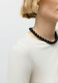 Persoon met kort blond haar, gekleed in een witte ribbed top en een chunky zwarte kralenketting, gedeeltelijk zichtbaar van schouder tot kin.