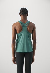 Persoon met gevlochten haar, gekleed in een groene sporttop zonder mouwen en zwarte leggings, staande met de rug naar de camera tegen een effen achtergrond.