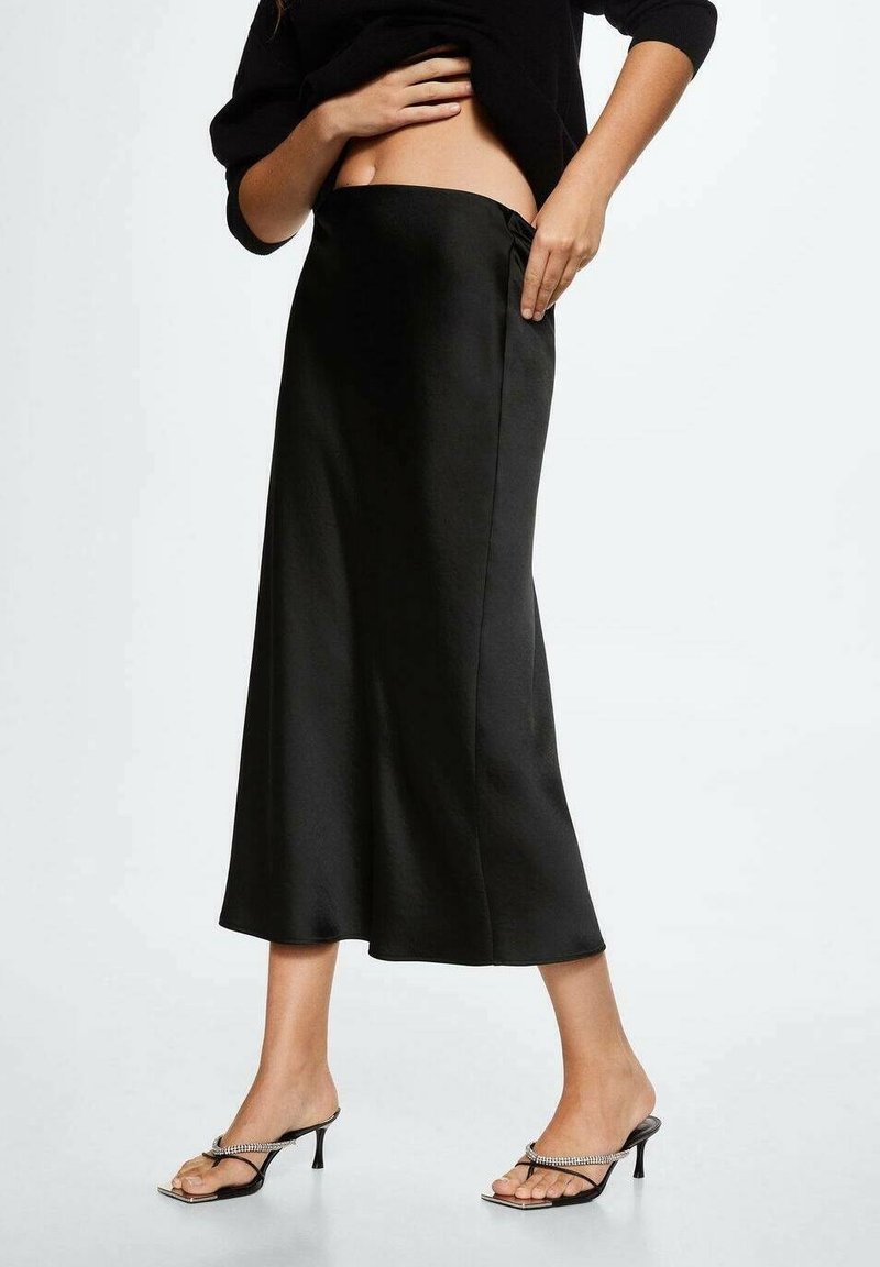 Femme ajustant une jupe midi en satin noir taille haute, portant un haut noir et des sandales à talons noires avec des brides ornées de strass sur fond clair.