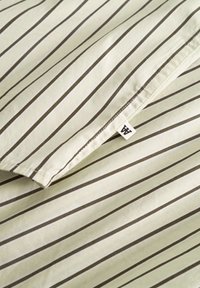 Țesătură în dungi de culoare galben pal, cu linii verticale subțiri de culoare maro închis. Include o mică etichetă albă cu inițiale în colț.