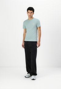 Lysegrøn bomuld T-shirt med et lille logo på brystet, parret med sorte løstsiddende bukser og grå sneakers. Enkel og afslappet outfit.