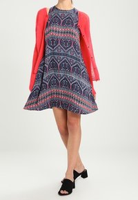 Navy Blaues Kleid mit buntem Paisley-Muster, ausgestelltem Rock und pinker Strickjacke. Schwarze offene Absatzsandalen.