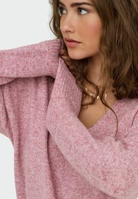 Jeune femme aux cheveux bruns ondulés portant un pull en laine rose doux et un collier en or avec de petites pendules en forme de diamant.