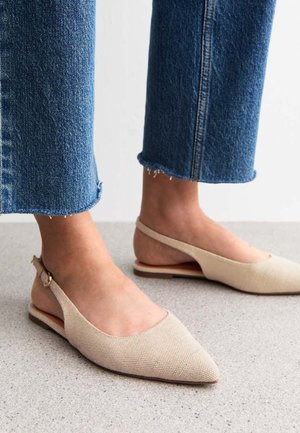 Füße, die beige spitze Slingback-Ballerinas tragen, kombiniert mit gekürzten blauen Jeans auf einem hellgrauen Boden.