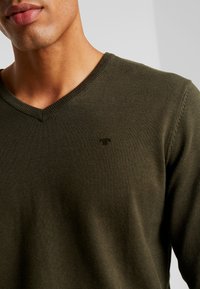 Olivgrüner, mittelschwerer Strickpullover mit V-Ausschnitt, ausgestattet mit einem subtilen, tonalen Logo in der Nähe der Brust sowie ribbelndem Nacken- und Ärmelsaum.