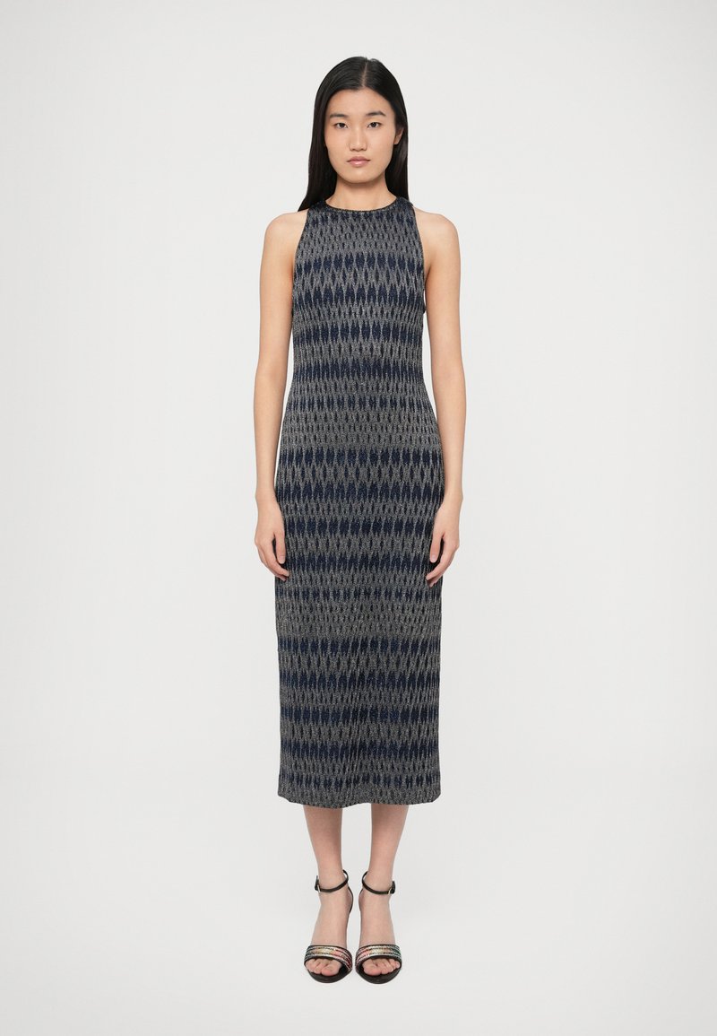 Moteris stovinti, dėvinti be rankovių, raštuotą tamsiai mėlynos ir pilkos spalvų midi suknelę su kulniukais su čiurnos dirželiais, neryškioje fone.