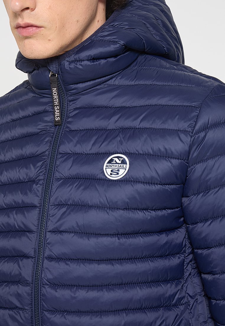 Mężczyzna w granatowej pikowanej kurtce z kapturem, z logo North Sails na klatce piersiowej i markowym suwakiem, zwrócony w lewo.
