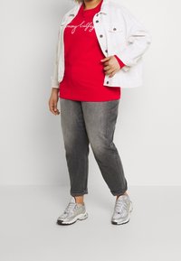 Rode sweatshirt met wit logo, gecombineerd met een witte denimjack, grijze tapered jeans en zilveren sport schoenen.