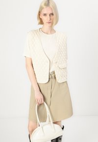 Beige quiltet vest med lommer, brukt over en hvit stripete topp, sammen med en khaki skjørt. Holder en hvit håndveske med buede håndtak.