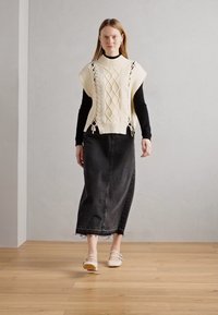 Stickad krämfärgad väst med kabelmönster över en svart långärmad skjorta, i kombination med en mörk denimmaxikjol och ljusa slip-on-skor.