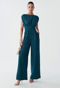 Woman in a teal jumpsuit with wide legs, holding a gold woven handbag, standing confidently against a light background.
