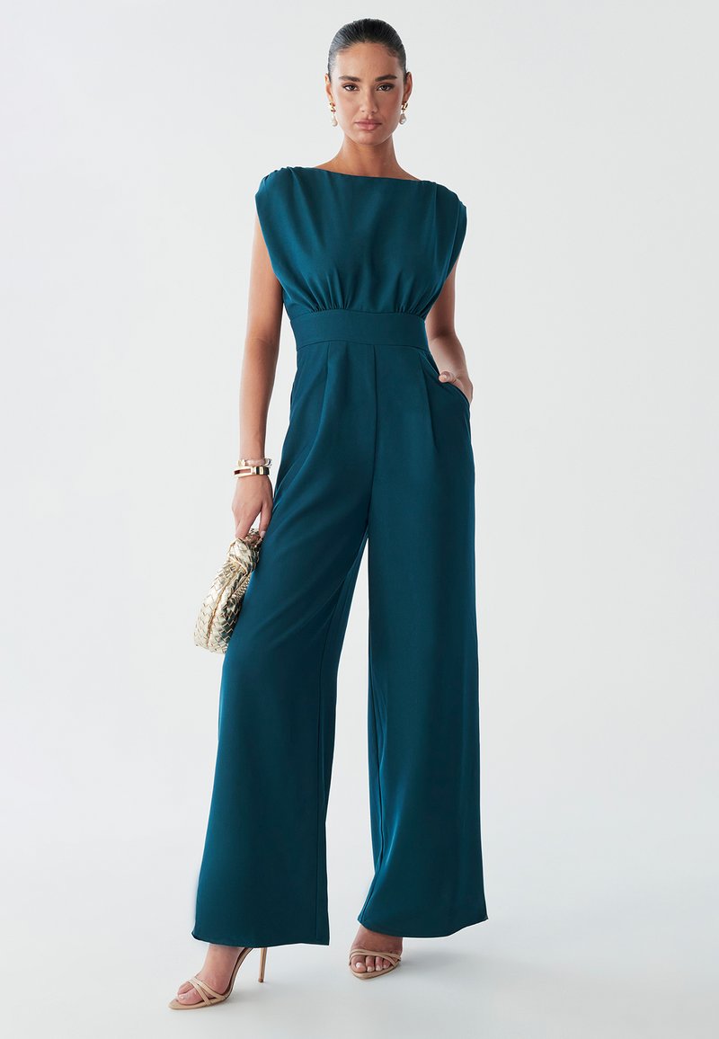 Woman in a teal jumpsuit with wide legs, holding a gold woven handbag, standing confidently against a light background.