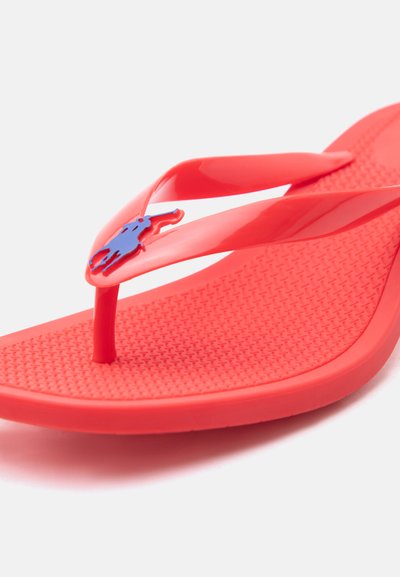 Φωτεινό κόκκινο σαγιονάρι με υφή στη σόλα και μπλε λογότυπο αλόγου στο λουράκι, εμφανίζεται σε λευκό φόντο.