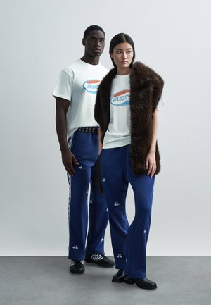 Λευκά γραφικά μπλουζάκια με το λογότυπο "SEX", μπλε φόρμες με κεντημένα σχέδια και καφέ πιλημένα γούνινα σάλι. Κανονική, άνετη εφαρμογή.