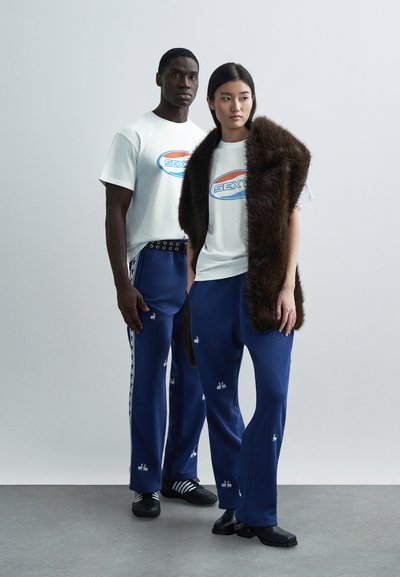 Vita grafiska t-shirts med "SEX"-logotyp, blå sweatpants med broderade motiv och en brun fuskpäls-sjal. Avslappnad och ledig passform.