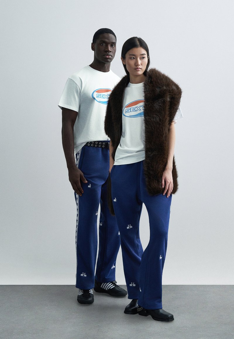 Bijele grafičke majice s logotipom "SEX", plave trenirka hlače s vezom i smeđi šal od umjetnog krzna. Ležerni, opušteni kroj.