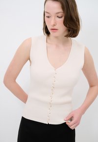 Beige Ripp-Top mit V-Ausschnitt, ausgestattet mit einer Reihe kleiner, runder Knöpfe an der Vorderseite. Hergestellt aus einem weichen, strukturierten Material.
