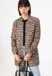Jachetă cu imprimeu leopard, cu guler, interior negru, mâneci lungi și croială lejeră, asortată cu blugi de culoare albastru deschis și o geantă neagră.