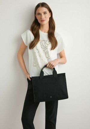 Kobieta o długich brązowych włosach w białej koszuli ze złotym wzorem, czarnych spodniach, trzymająca dużą czarną torbę typu tote na tle białej ściany.