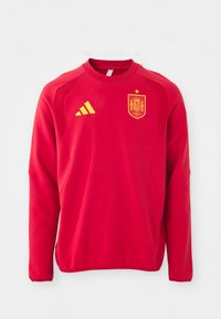 Sarkans džemperis ar garām piedurknēm, apaļu kaklu, teksturētām plecu daļām, dzeltenu Adidas logotipu un dzeltenu Spānijas nacionālās komandas emblēmu.