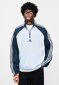 Ανοιχτό μπλε και ναυτικό αμάνικο αντιανεμικό με λευκές ρίγες. Διαθέτει ψηλό γιακά, φερμουάρ στο μισό εμπρός μέρος και το λογότυπο της Adidas στο στήθος.