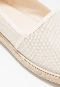 Beige-kangast slip-on-kengät, joissa on teksturoitu päällinen, viimeistelty kudotulla jute-pohjalla ja sileällä kumireunalla. Minimalistinen muotoilu.