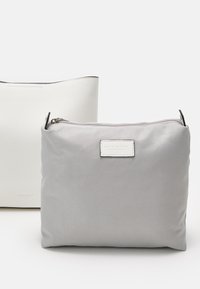 Twee tassen: een grijze stoffen pouch met een ritssluiting en een witte leren tas met structuur en een gladde bovenrand, voorzien van logo-accenten.