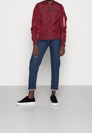 Personne portant une veste bomber rouge, un jean bleu retroussé et des baskets noires à scratch, debout devant un fond blanc uni.