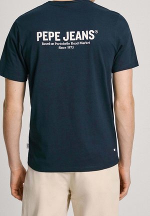 Osoba oblečená v námořnicky modrém tričku Pepe Jeans s bílým textem na zadní straně a béžových šortkách, viděná zezadu.
