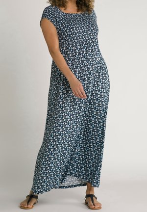 Kvinde iført en ankellang mørkeblå kjole med hvidt blomsterprint og sorte T-remssandaler, stående på en ensfarvet baggrund.