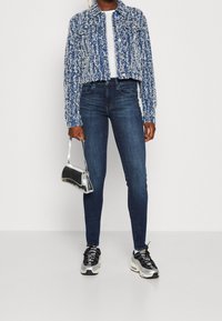 Blå och vit texturerad kortjacka över en vit skjorta, matchad med mörkblå skinny jeans och silverfärgade sneakers, hållande en silverhandväska.