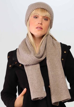 Beige knit scarf with ribbed texture and chevron accents, paired with a matching beanie, worn over a black jacket.