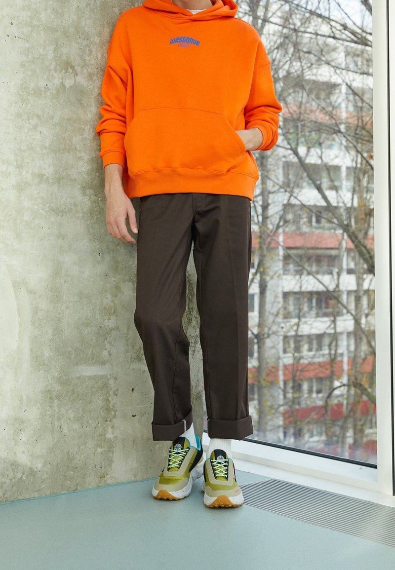 Sudadera naranja brillante con bolsillo frontal y logotipo azul, combinada con pantalones marrones y zapatillas multicolores con suelas blancas.