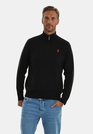Mann trägt einen schwarzen Quarter-Zip-Pullover mit rotem Logo und hellblauen Jeans, steht mit einer Hand in der Tasche vor einem weißen Hintergrund.