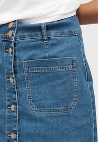 Jupe en denim bleu taille haute avec boutons à l'avant, une poche arrière unique, et un haut blanc rentré, vue de la taille vers le bas.