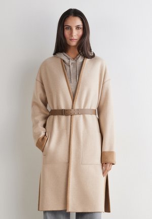 Femme portant un manteau beige ceinturé ouvert sur un sweat à capuche gris, debout contre un mur clair uni, une main dans la poche.