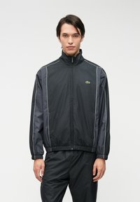 Μαύρο αδιάβροχο μπουφάν με ψηλό γιακά, ελαστικούς καρπούς και στρίφωμα. Διαθέτει ανακλαστικές λευκές ρίγες κα ι πράσινο λογότυπο στο στήθος.