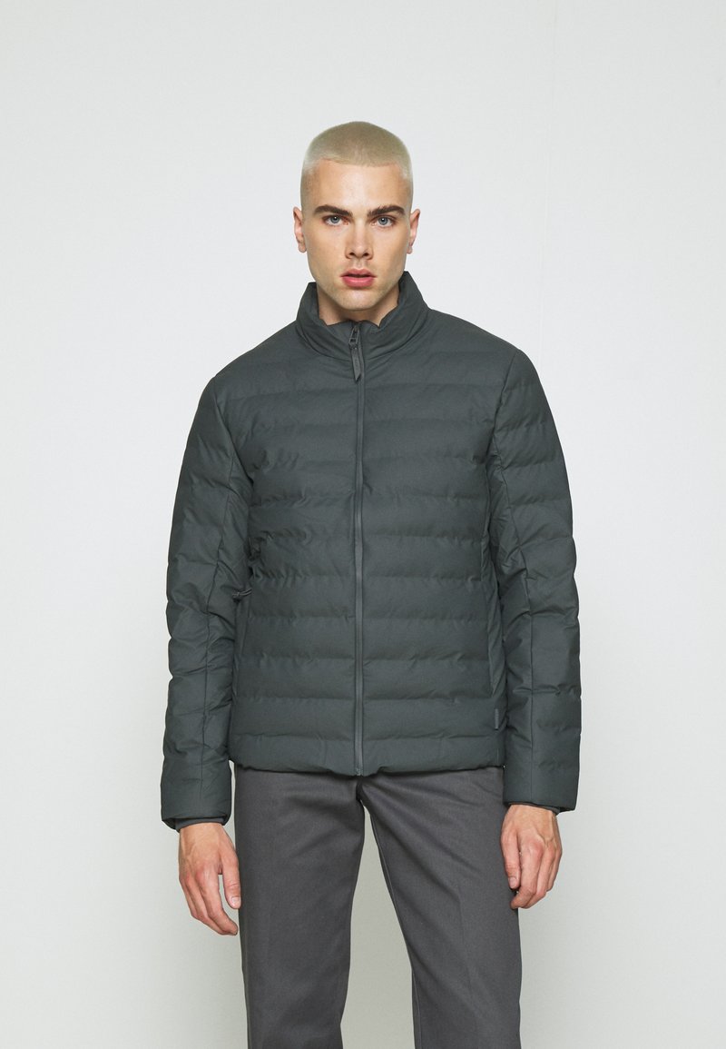 Veste doudoune gris foncé avec un design matelassé horizontal, col haut et fermeture éclair sur le devant. Assortie avec un pantalon gris, le modèle se tient devant un fond clair.