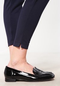 Black patent leather loafers with a flat sole and decorative tassel detail, worn with navy cropped pants featuring slit hems.