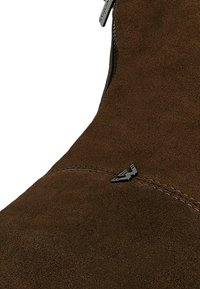 Bruine suède enkellaars met een ritssluiting en een zwart metalen logo-accent. Het ontwerp bevat gladde stiknaden langs de naad.