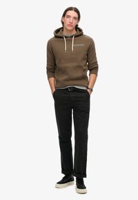 Sudadera marrón con cordones y logo, combinada con pantalones negros y zapatillas negras con suela blanca. Textura suave, corte estándar.