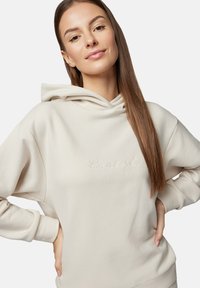 Jasno beżowa bluza z kapturem wykonana z miękkiego materiału. Posiada sznurki w kapturze oraz wyhaftowaną nazwę marki w subtelnej czcionce na przedniej części.