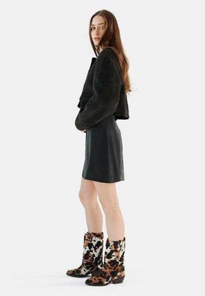 Veste noire texturée courte, jupe en cuir noir cintrée, et bottes à imprimé vache marron et blanc à hauteur de genou avec des talons épais.