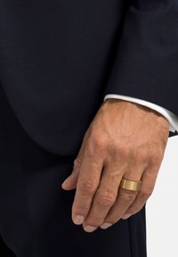 Anello in metallo dorato su una mano con pelle chiara, indossando un abito scuro con una camicia a colletto bianco visibile al polsino. Texture liscia, forma rettangolare.